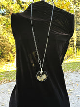 Load image into Gallery viewer, Long necklace with a circular pendant on a black dress against a blurred outdoor background
